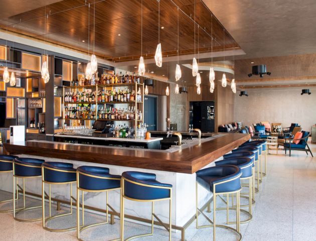 This image shows a modern bar with blue stools, a well-stocked liquor shelf, overhead pendant lights, and a contemporary seating area in the background.