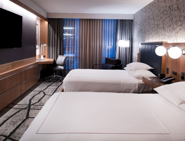 A modern hotel room with two beds, wall-mounted TV, desk, chair, and bedside lamps, featuring contemporary decor and large windows.