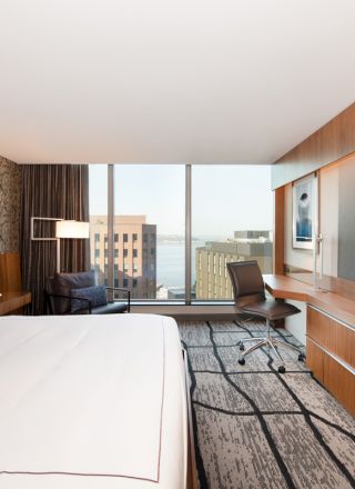 A modern hotel room with a king-sized bed, large TV, desk, and chair. The room features a window with a city and water view in the background.