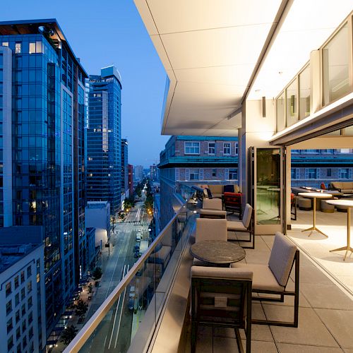 An image showing a modern outdoor terrace with seating, overlooking a cityscape with high-rise buildings at dusk, providing a scenic urban view.