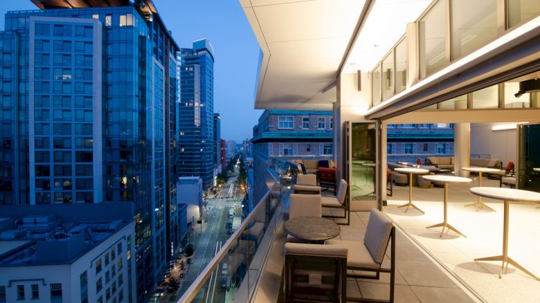 An evening cityscape view from a modern balcony with tables, chairs, and glass railings; adjacent buildings and a street are visible.