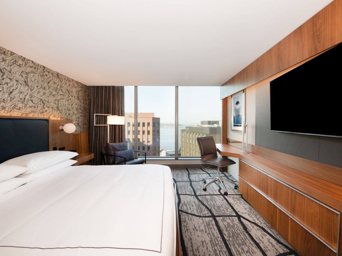 A modern hotel room features a king-sized bed, a large flat-screen TV, a work desk, a chair, and a window offering a city view with buildings visible.
