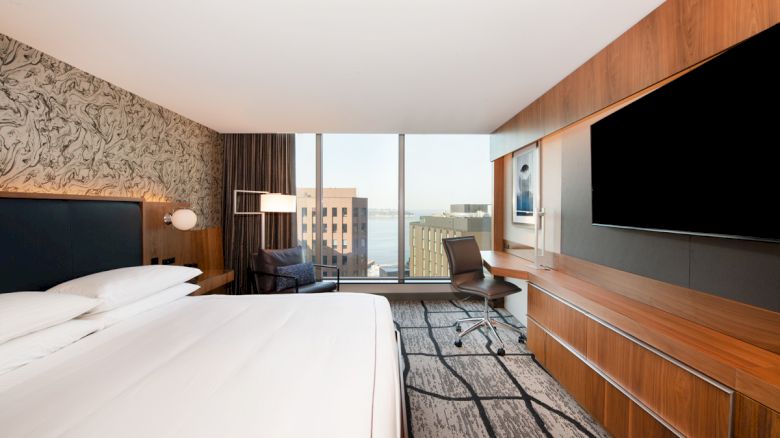 A modern hotel room features a king-sized bed, a large flat-screen TV, a work desk, a chair, and a window offering a city view with buildings visible.