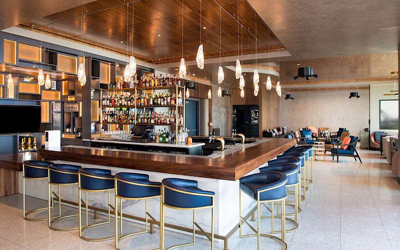 A modern bar with blue cushioned chairs, pendant lighting, and a well-stocked bar counter in an elegant, minimalist setting.
