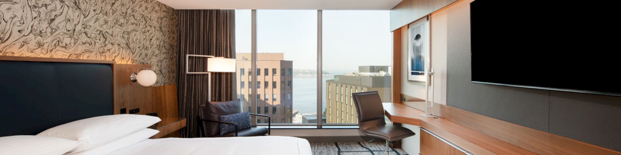 This image shows a modern hotel room with a large bed, a desk, a flat-screen TV, and a view of city buildings through a large window.